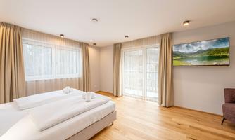 A modern bedroom with two single beds, large windows, and wooden flooring. A television hangs on the wall, and there is a cozy seating area.