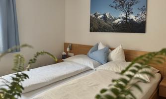 A cozy bedroom with two beds and a beautiful view of the mountains. The room features plants and gentle lighting conditions.