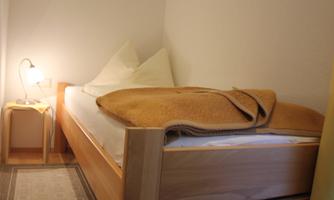 A cozy bedroom with a single bed and a warm blanket. Next to the bed is a lamp on a small table.