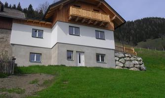 A modern house with a wooden railing on the balcony stands in a green meadow. In the background, wooded hills can be seen.