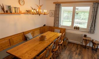 A cozy dining area with a large wooden table and chairs. On the wall, there is a shelf with decorations and books.