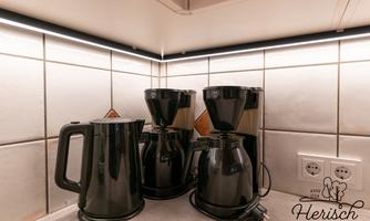 Two black coffee machines and a kettle are standing on a countertop. The environment is bright and modern, with tiles and indirect lighting.