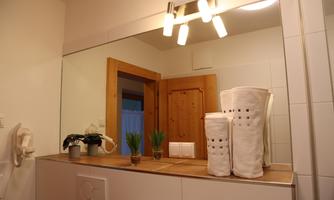 A modern bathroom with a large mirror and warm lighting. On the shelf, there are plants and clean towels.