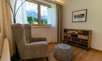 A cozy living space with a gray armchair and a round stool. Through the window, there is a beautiful view of the mountains and the green landscape.