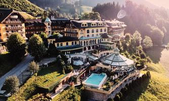 An elegant hotel building on a hill, surrounded by green forests and mountains. In the foreground, there is a swimming pool and a glass veranda.