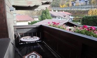 A cozy balcony with a small table and blooming plants. In the background, houses and green hills can be seen.