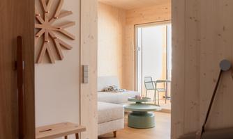 A modern room with wooden walls and soft lighting. In the background, there is a bed and a small table with chairs.
