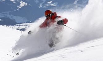 A skier navigates through fresh powder snow in the mountains. The landscape is clear and wintry, with snow-covered peaks in the background.
