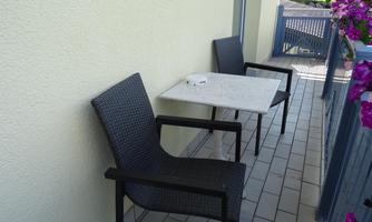 A small balcony with two black chairs and a table. To the right, colorful flowers can be seen on the railing.