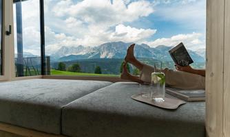 A cozy sitting area with a view of mountains and a blue sky. A person is relaxing with a book and a drink.
