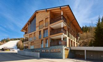 A modern wooden building with multiple balconies and large windows. In the background, a ski slope and a green forest are visible.