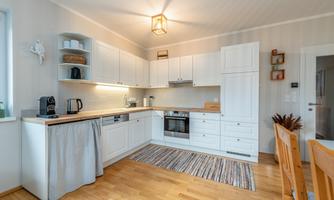 A modern kitchen with white cabinets and a wooden table. The space is bright and inviting, with a cozy rug and some decorative elements.