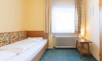A simple room with a single bed, a table, and a window. The walls are in soft colors, and the floor has a carpet.