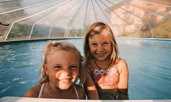 Two cheerful girls are standing in a pool and smiling at the camera. The surroundings are bright and friendly, with a roof over the water.