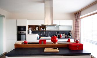 A modern kitchen with white cabinets and black countertops. Red accents and a large window front create a bright atmosphere.