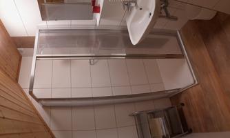 A small bathroom with a shower and a sink. It has white tiles and a wooden floor.