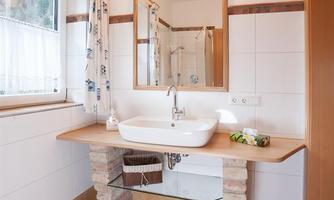 A modern bathroom with a simple sink and a large mirror. The walls are bright, and there are windows that provide ample light.