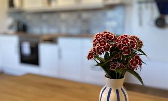 An elegant vase with red flowers stands on a wooden table. In the background, a modern kitchen can be seen.