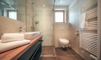 A modern bathroom with a glass shower cubicle and a sink. The walls are light tiled and there are fresh towels on the shelf.