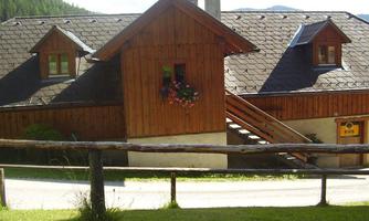 A cozy wooden house with a balcony and colorful flowers. The surroundings are green and hilly, ideal for a peaceful retreat.