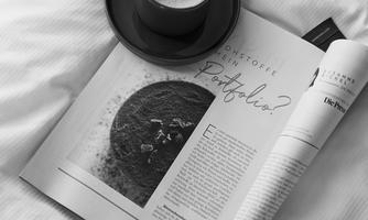A cup of coffee is on an open magazine lying on a bed. The atmosphere is cozy and inviting.