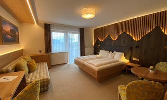 A modern hotel room with a double bed, a cozy sofa, and a table. Large windows provide plenty of daylight and a view outside.