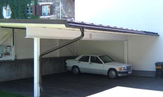 A white car is parked in a covered garage. A building is visible in the background.