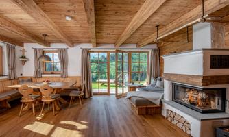 A cozy living room with wooden floors and large windows. In the background, you can see a fireplace and a dining area with wooden chairs.