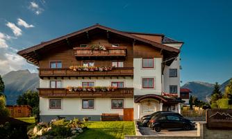 A cozy building in the mountains with balconies full of flowers. In the foreground, there is a black car on a green meadow.