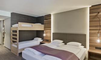A modern hotel room with a double bed and a bunk bed. The walls are painted in neutral colors and the lighting is stylish.