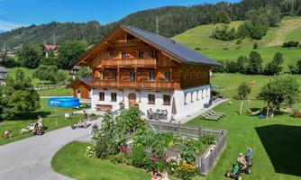 A traditional wooden house with a balcony in a rural setting. Children are playing in the garden and the landscape is green and hilly.