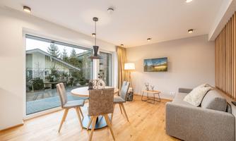 A modern furnished living area with a round dining table and chairs. Large windows let in plenty of daylight and offer a view of the outdoor facilities.