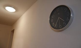 A wall clock with a black dial hangs on a light-colored wall. Above it is a ceiling lamp that illuminates the room.