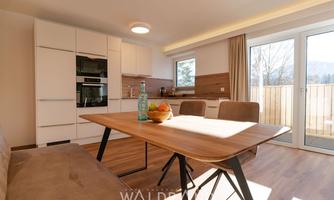 A modern kitchen with a dining area and large windows. The table is decorated with fruit and a bottle, creating an inviting atmosphere.