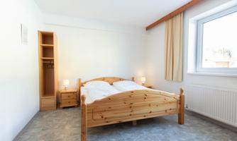 A cozy bedroom with a wooden double bed. There is a wardrobe and plenty of natural light coming through the window.