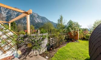 A beautiful garden with green lawn and blooming plants. In the background, impressive mountains and a clear blue sky can be seen.