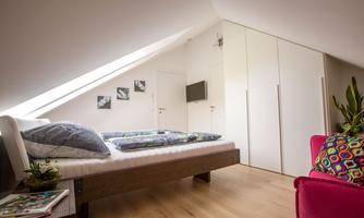 A modern bedroom with a sloped ceiling and wooden floor. The furniture is minimalist and bright, with a colorful seating area.