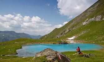 A picturesque mountain lake with bright blue water, surrounded by green meadows and high mountains. A person sits by the shore and enjoys nature.