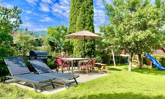 A beautiful garden with loungers, a table and chairs under a sunshade. There are lush plants and a playground with a slide.
