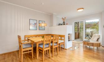 A modern dining area with a wooden table and chairs. Large windows provide plenty of natural light and a view of the balcony.