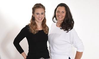 Two women are smiling in front of a bright background. The younger one is wearing a black top, and the older one is wearing a white one.