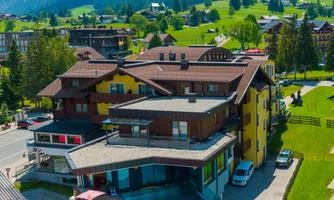 A modern hotel with a swimming pool and sun loungers. The environment is characterized by green meadows and mountains.