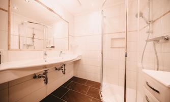 A modern bathroom with a shower, a double sink, and bright walls. The floor is laid with dark tiles, and there is a large mirror.