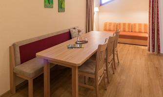 A bright dining room with a long wooden table and several chairs. Two pictures hang on the wall, and a cozy couch is placed on the side.