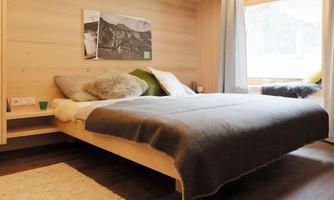 A modern bedroom with a simple bed and cozy pillows. Large windows allow plenty of daylight in and provide a view outside.