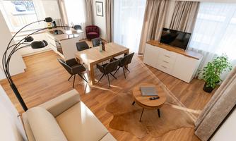 A modern living room with a dining area and wooden floor. Large windows with curtains provide lots of daylight.