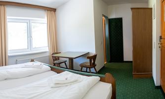A simple room with two beds, a table, and chairs. The room has large windows and a bright, friendly impression.