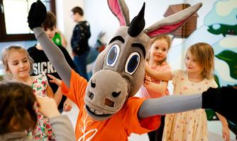 A donkey mascot in an orange T-shirt is hugging happy children. The children are laughing and playing in a colorful room.