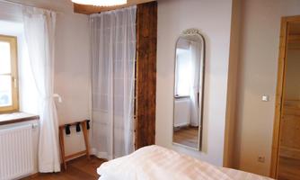 A cozy bedroom with wooden details and large windows. The room has a large mirror and fresh white curtains.