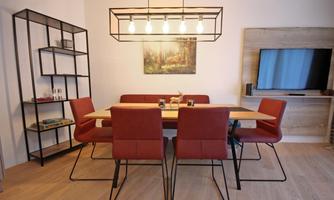 A modern dining area with a large table and red chairs. In the background, there is a shelf and a television.